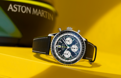 The Breitling Navitimer B01 Chronograph Aston Martin Edition is displayed at an angle on a yellow pedestal; the Aston Martin branding can be seen blurred in the background.