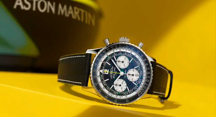 The Breitling Navitimer B01 Chronograph Aston Martin Edition is displayed at an angle on a yellow pedestal; the Aston Martin branding can be seen blurred in the background.