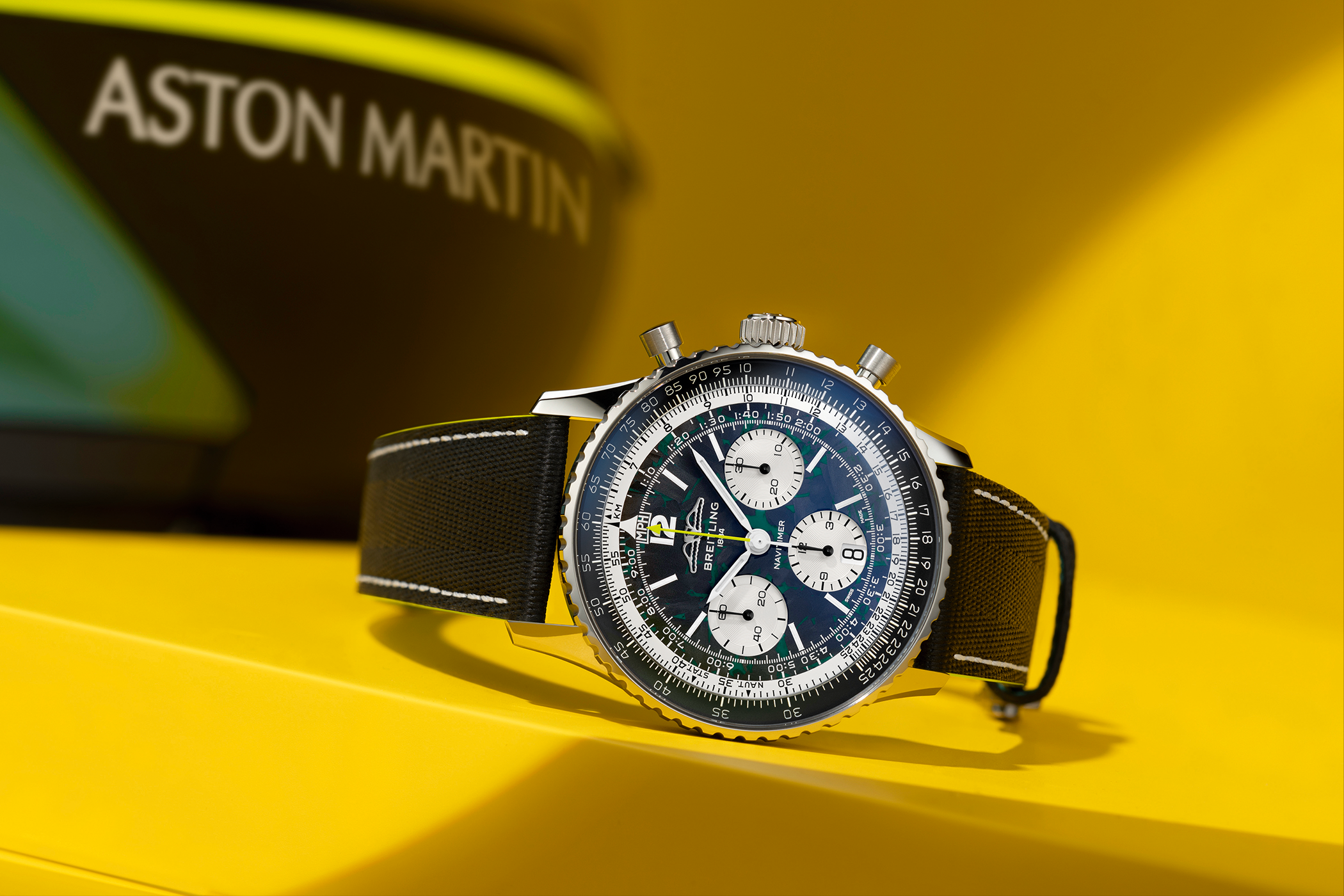 The Breitling Navitimer B01 Chronograph Aston Martin Edition is displayed at an angle on a yellow pedestal; the Aston Martin branding can be seen blurred in the background.